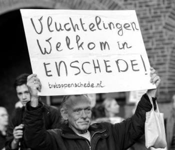 A black and white photo of a man holding a sign written in Dutch that reads Refugees Welcome in Enschede