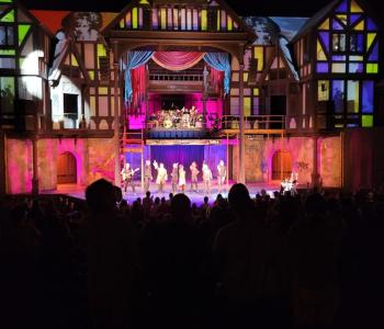 Photo of people on stage with the audience darkened in the foreground and a lit exterior of buildings in the background.