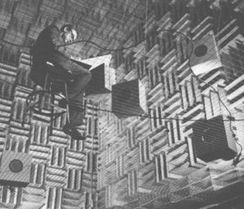 A scientist takes notes while sitting in a chair while insdie the Murray Hill Anechoic Chamber, which has hanging speakers and walls made of grids. 