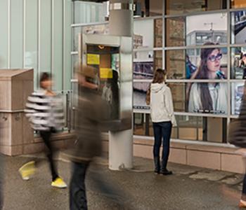 Five people walk by each other, blurred by their motion while one person is still looking at her reflection in a bank of monitors. 